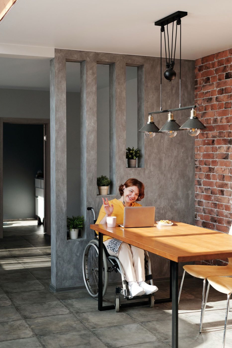 A woman in a wheelchair, smiling, waving on a video call at her laptop at a home dining table.