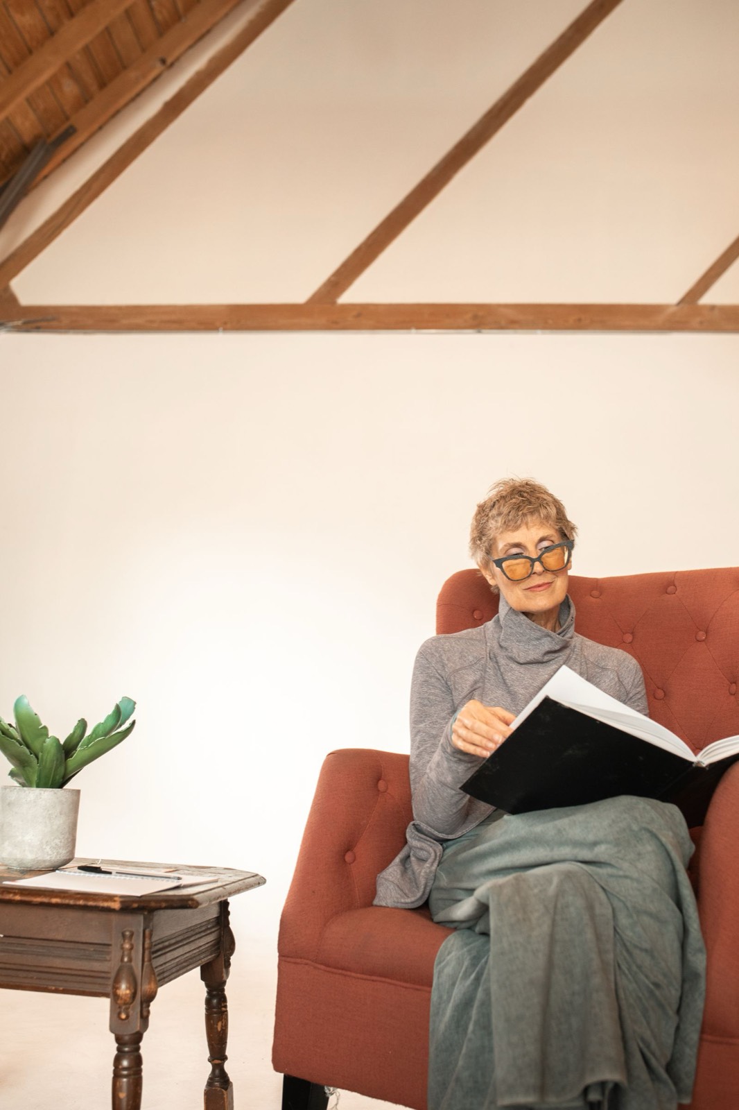 An older woman reading a book in an armchair, sunglasses on, quiet studio light.