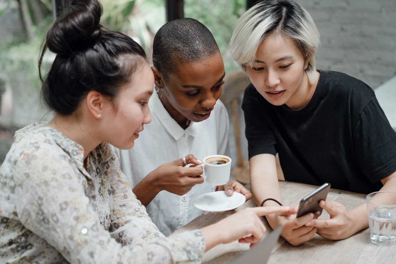 Three women leaning in together over a phone at a caf&eacute; table, one holding a coffee cup.