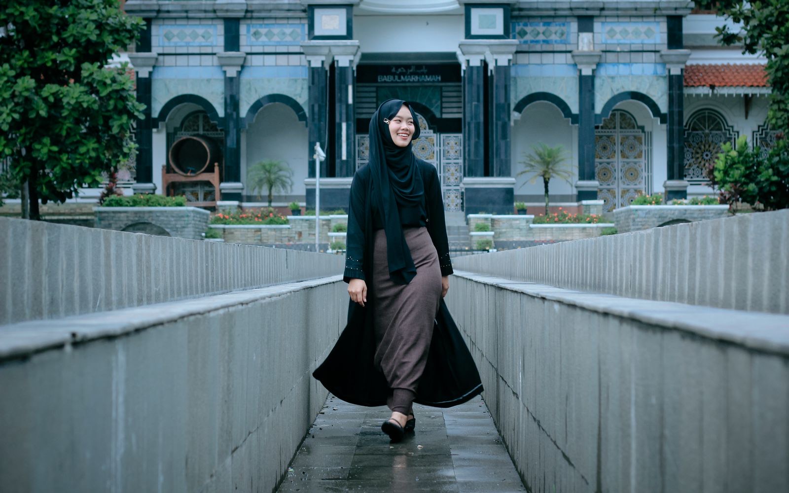 A woman in her twenties in a headscarf, smiling, walking through a sunlit architectural courtyard.